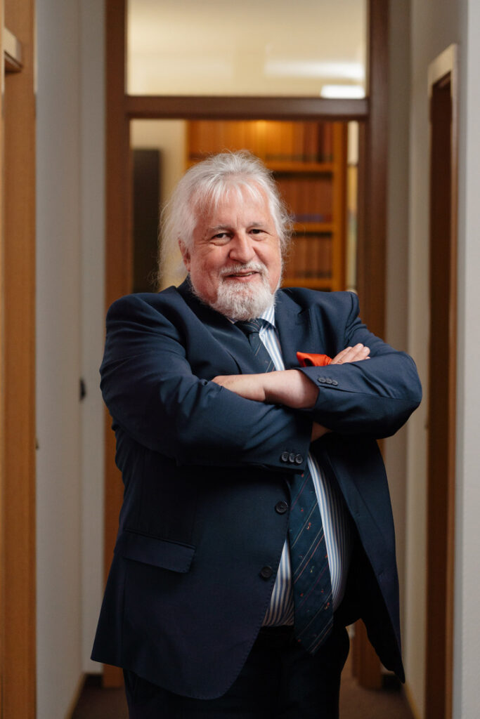 Porträt eines erfahrenen Geschäftsmanns mit grauen Haaren und Bart im Anzug und gestreifter Krawatte, verschränkte Arme im Büroflur