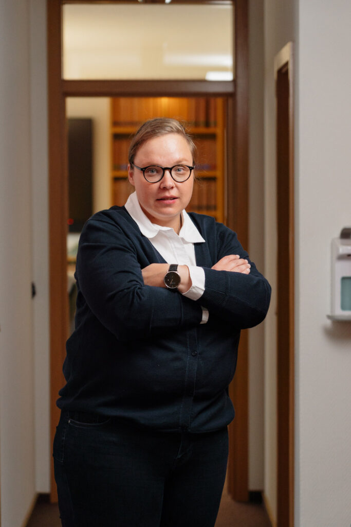 Porträt einer selbstbewussten Frau mit Brille und dunkler Kleidung, verschränkte Arme im Büroflur