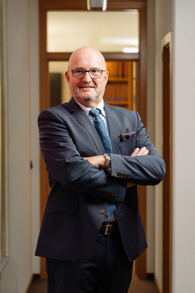 Porträt eines lächelnden Geschäftsmanns mit Brille im dunklen Anzug und blauer Krawatte, verschränkte Arme im modernen Büroflur