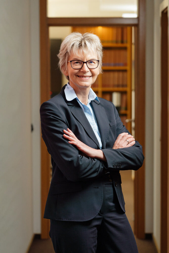 Porträt einer lächelnden Frau mit Kurzhaarfrisur und Brille im dunklen Anzug und hellblauer Bluse, verschränkte Arme im modernen Büroflur