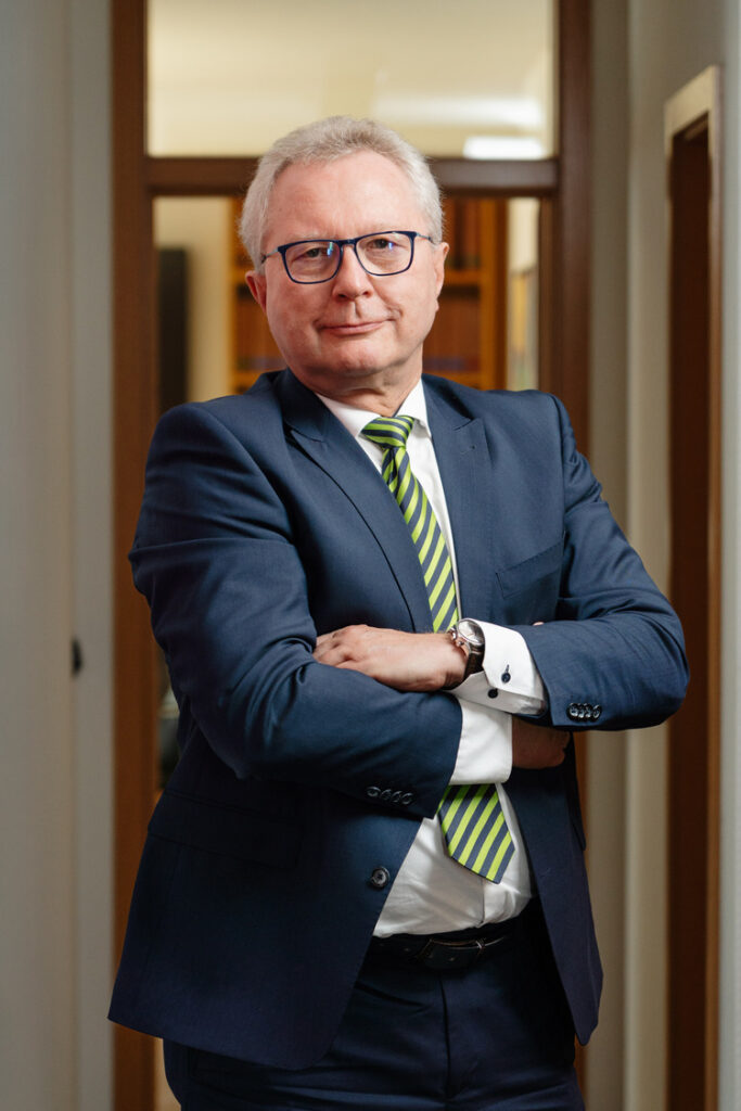 Porträt eines Geschäftsmanns mit Brille im dunklen Anzug und grün gestreifter Krawatte, verschränkte Arme im modernen Büroflur