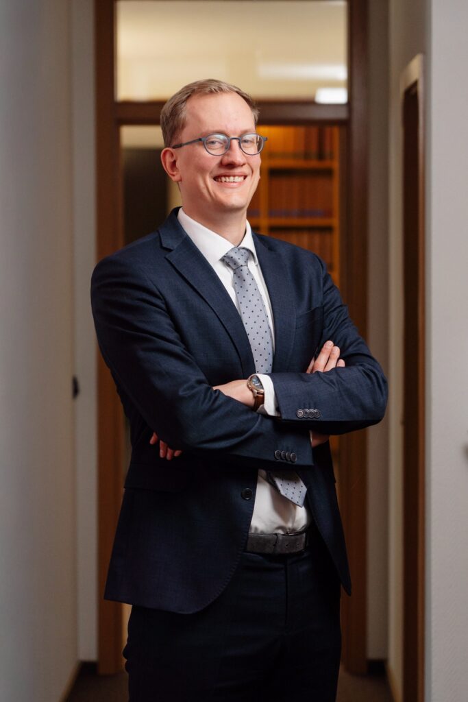 Ein junger Anwalt der Kanzlei Kessing • Hespe • Dr. Steenken lächelt freundlich in die Kamera, während er seine Arme verschränkt.